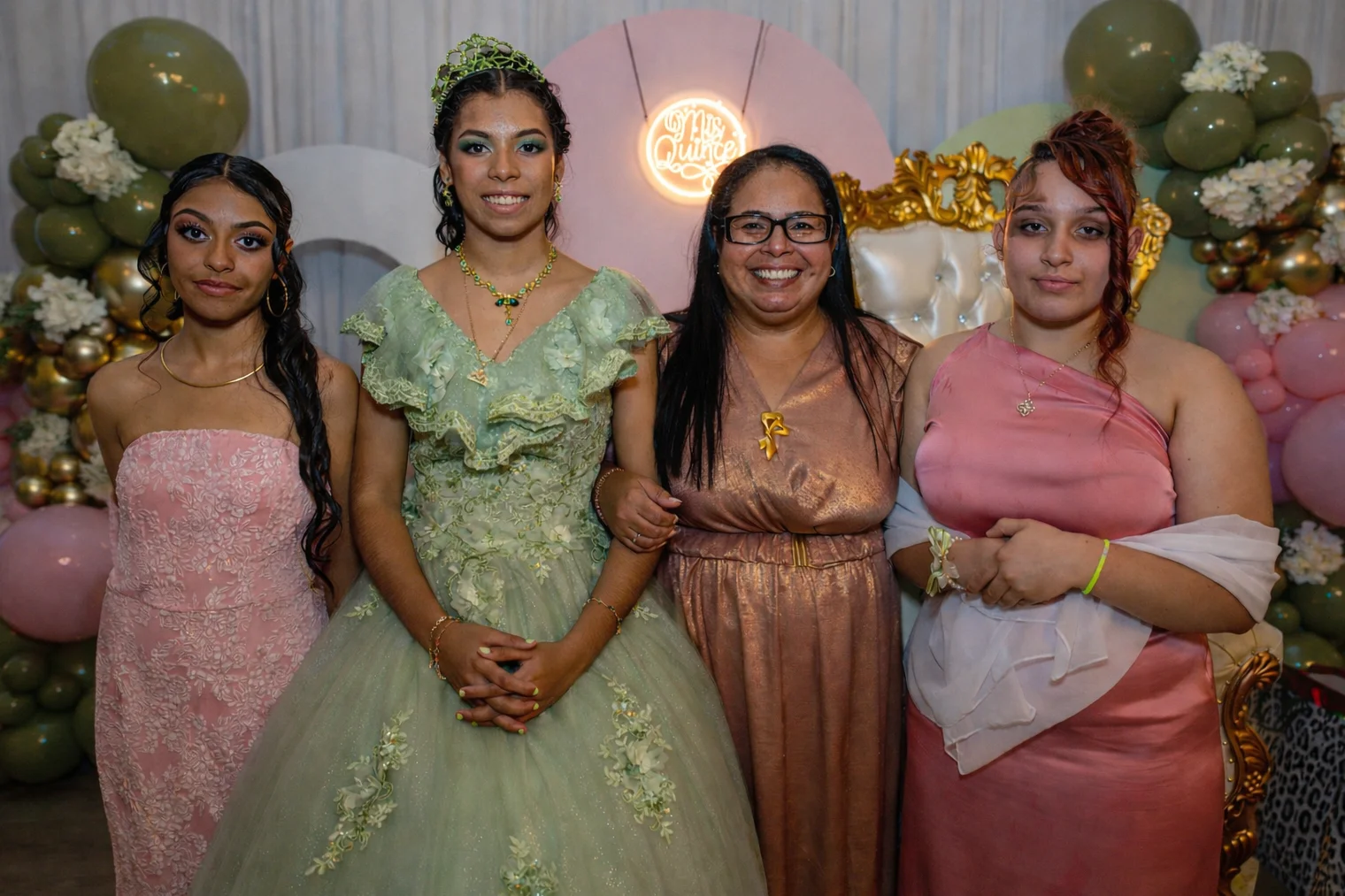 A quinceañera celebrant in a green gown posing with three guests beside balloon decorations.