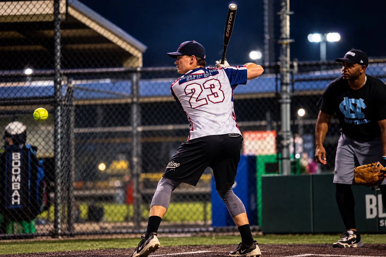 A softball batter swinging as the ball approaches at night.