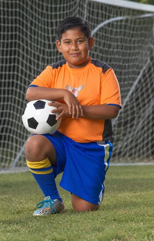 A young soccer player kneeling with a ball in front of a goal.